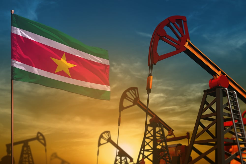 Suriname oil industry concept, industrial illustration. Fluttering Suriname flag and oil wells on the blue and yellow sunset sky background - 3D illustration